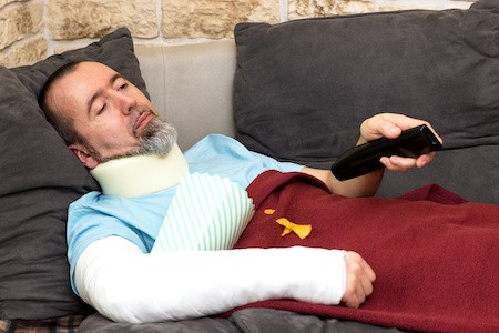 MoneyCheck | Arbeitsunfähigkeitsversicherung, Mann liegt verletzt auf dem Sofa MoneyCheck | Arbeitsunfähigkeitsversicherung